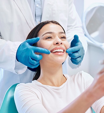 Dentist touching corner of woman’s mouth as she smiles into handheld mirror